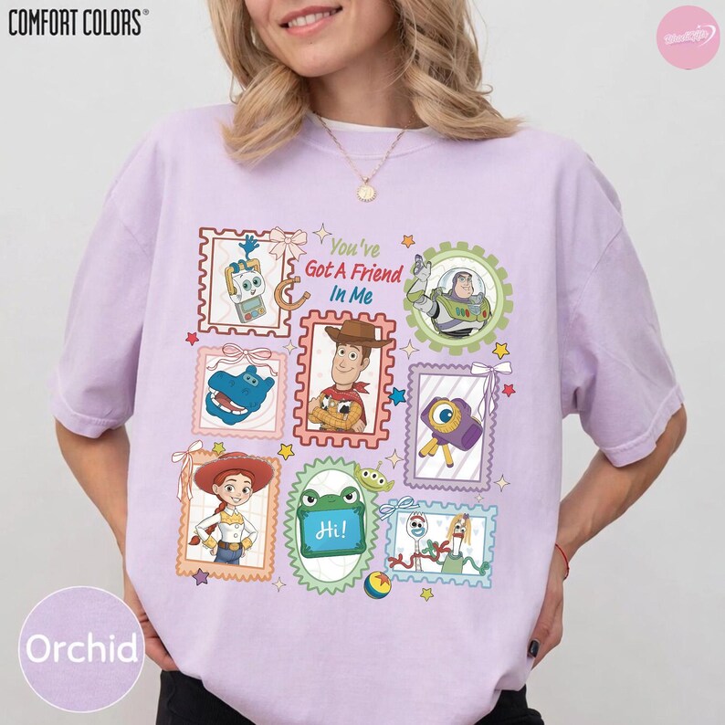 May include: A light purple t-shirt featuring a graphic print of various characters from the Toy Story franchise arranged in postage stamp-style frames. The text "You've Got A Friend In Me" is printed in the center, surrounded by colorful illustrations.