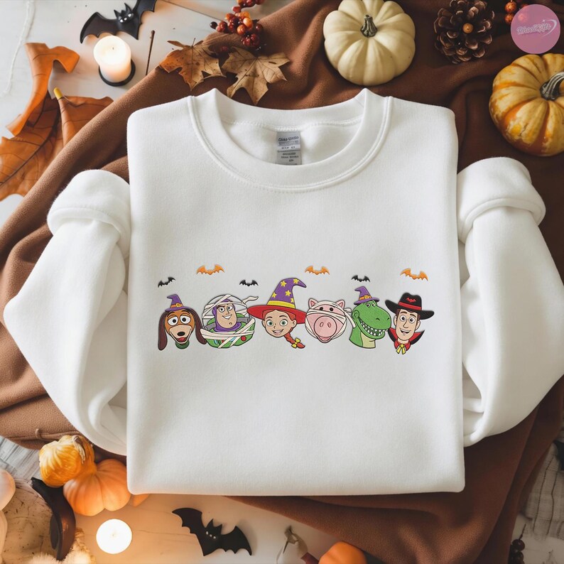 May include: White crewneck sweatshirt featuring a colorful cartoon graphic of Toy Story characters dressed in Halloween costumes. The design includes characters like Woody, Buzz Lightyear, and others, with bats and pumpkins.