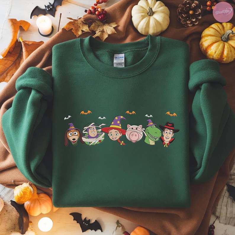 May include: A dark green sweatshirt featuring a Halloween-themed design with cartoon character faces. The design includes characters dressed in Halloween costumes, with bats above them. The sweatshirt is a Gildan brand.