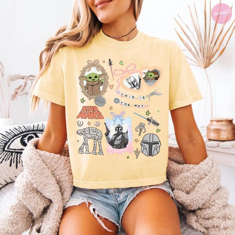 May include: Yellow t-shirt featuring Star Wars characters and imagery. The design includes illustrations of Baby Yoda, the Mandalorian, and the text "THIS IS THE WAY". The shirt has a crew neck and short sleeves.