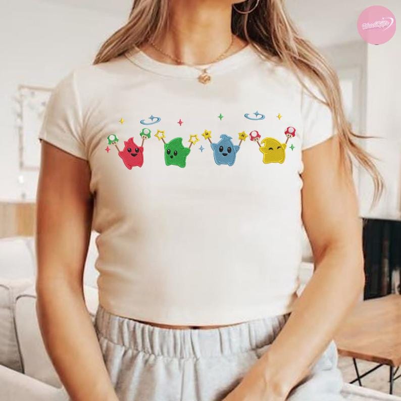 Puede incluir: Camiseta de color crema de manga corta con un dise&ntilde;o de dibujos animados colorido. El dise&ntilde;o incluye cuatro personajes en forma de estrella en rojo, verde, azul y amarillo, cada uno sosteniendo una estrella y una seta. La camiseta tiene cuello redondo.