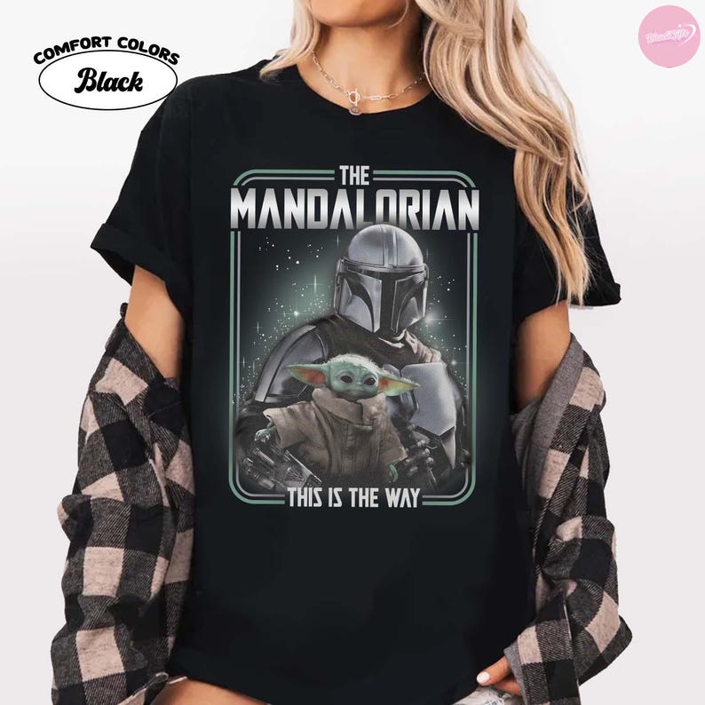 K&ouml;nnte beinhalten: Schwarzes T-Shirt mit einer Grafik von The Mandalorian und Grogu. Das Design enth&auml;lt den Text "THE MANDALORIAN" und "THIS IS THE WAY". Das Shirt ist mit "Comfort Colors Black" gekennzeichnet.
