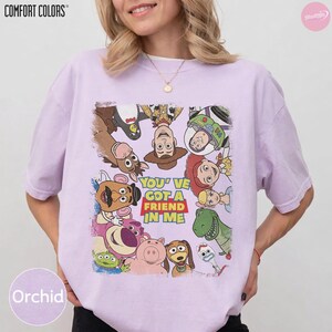 May include: Orchid Comfort Colors t-shirt featuring a graphic of characters from the movie Toy Story. The design includes Woody, Buzz Lightyear, and other characters with the text "YOU'VE GOT A FRIEND IN ME".