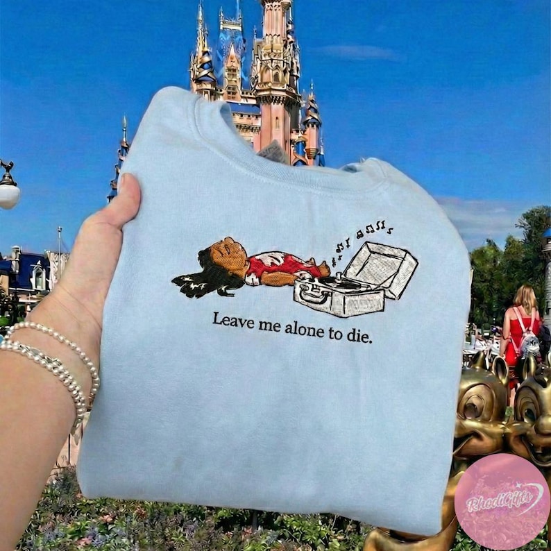 May include: Light blue sweatshirt with an embroidered graphic of a person lying next to a record player. The text "Leave me alone to die" is below the graphic. The background features a castle and blue sky.