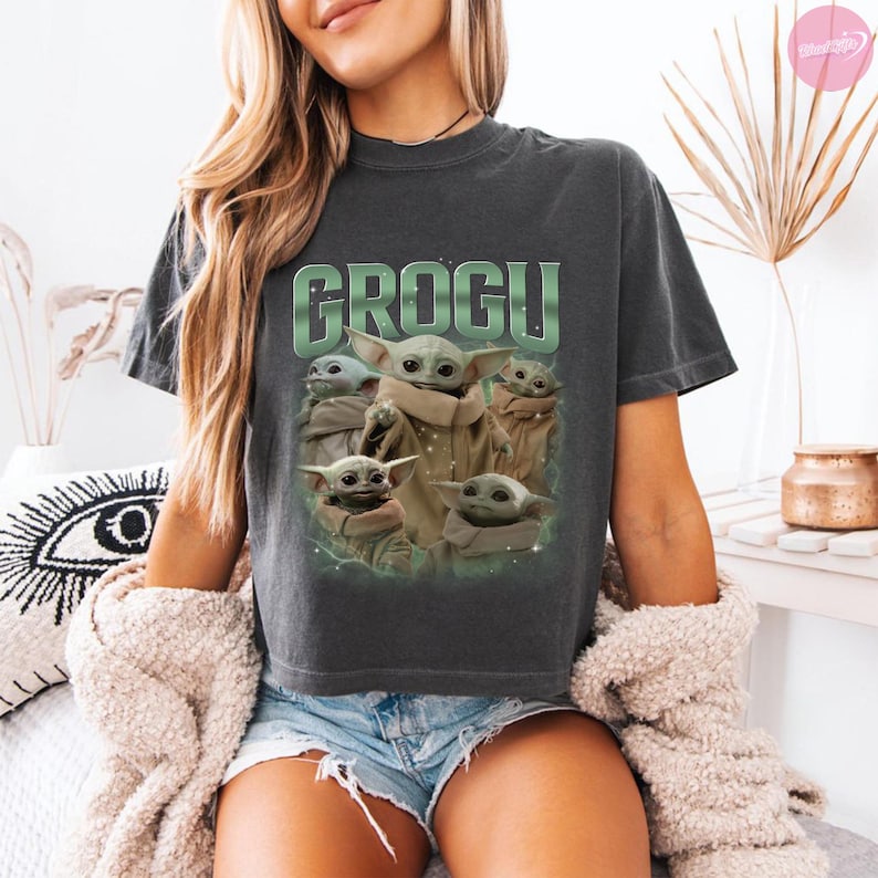 May include: Dark gray t-shirt featuring the word "GROGU" in green block letters above a graphic of Grogu from The Mandalorian. The graphic shows Grogu in various poses. The shirt is a crew neck.