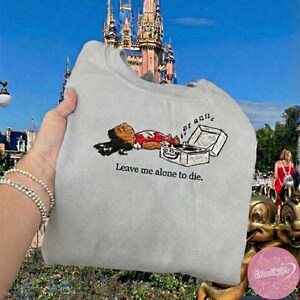 May include: A light gray sweatshirt featuring an embroidered cartoon character lying next to a record player. The text "Leave me alone to die" is printed below the image. The sweatshirt is held up in front of a castle.