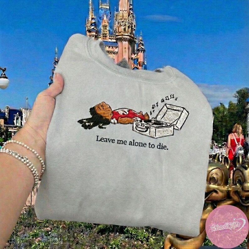 May include: A light gray sweatshirt featuring an embroidered cartoon character lying next to a record player. The text "Leave me alone to die" is printed below the image. The sweatshirt is held up in front of a castle.