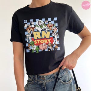 Puede incluir: Una camiseta negra de manga corta con un estampado gr&aacute;fico de varios personajes animados vestidos como profesionales m&eacute;dicos. El dise&ntilde;o incluye el texto "RN STORY" en letras grandes amarillas y rojas sobre un fondo de cuadros azul claro y blanco.