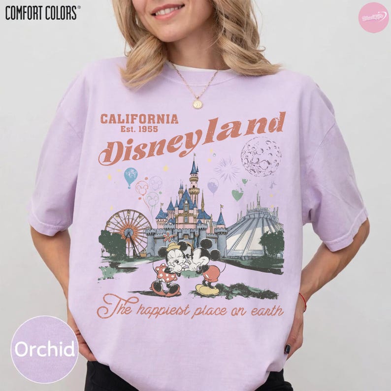 May include: Orchid-coloured t-shirt with a Disneyland graphic. The design features Mickey and Minnie Mouse, a castle, Ferris wheel, and the text "California Disneyland Est. 1955" and "The happiest place on earth."