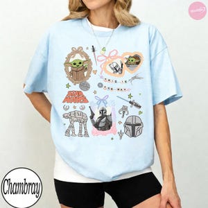 May include: Light blue t-shirt featuring various Star Wars-themed illustrations. The design includes images of Grogu, the Mandalorian, and the text "THIS IS THE WAY." Other graphics include the Star Wars logo, an AT-AT walker, and a Mandalorian helmet.
