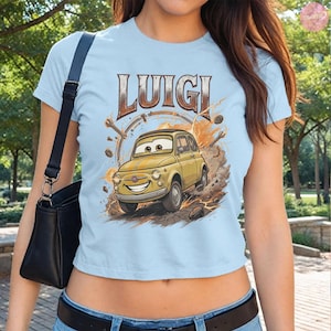 May include: Light blue t-shirt featuring the word "LUIGI" above a cartoon image of a yellow car. The car has a smiling face and is surrounded by tools and a brown background. The shirt is short-sleeved.
