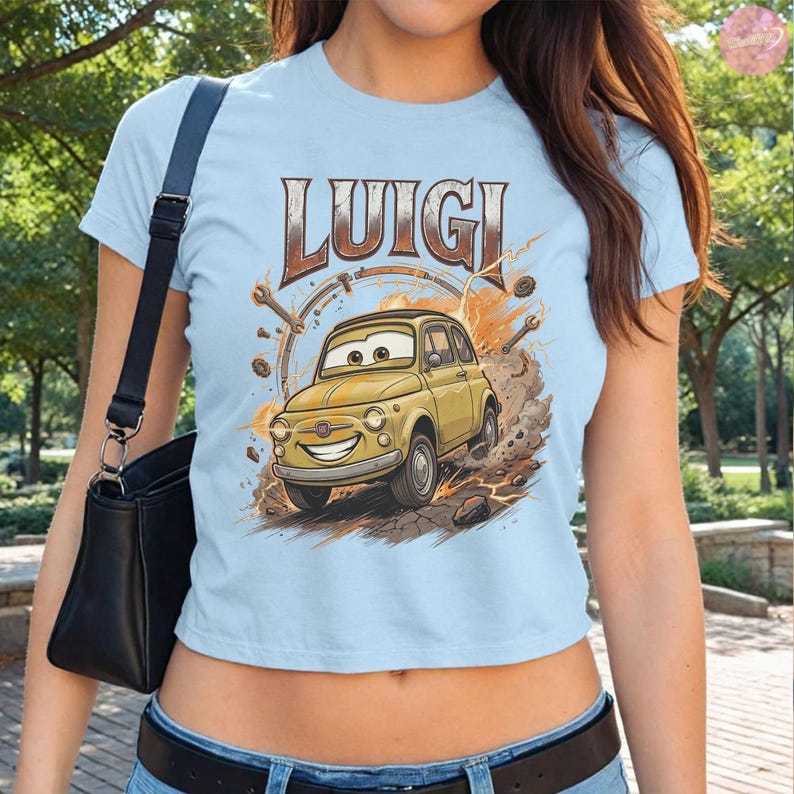 May include: Light blue t-shirt featuring the word "LUIGI" above a cartoon image of a yellow car. The car has a smiling face and is surrounded by tools and a brown background. The shirt is short-sleeved.