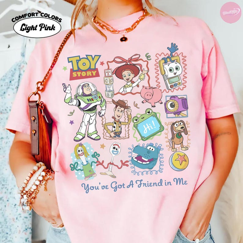 May include: A light pink t-shirt featuring a graphic print of Toy Story characters including Buzz Lightyear, Woody, Jessie, Hamm, and Forky. The design includes the Toy Story logo and the text You've Got A Friend in Me. The shirt is labeled Comfort Colors.