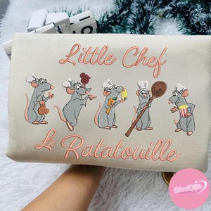 Pode incluir: Almofada retangular bege com texto bordado "Little Chef La Ratatouille". Apresenta cinco ratos chef de desenho animado em v&aacute;rias poses, segurando comida e uma colher de pau. O texto e os contornos dos ratos s&atilde;o rosa.