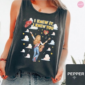 May include: A charcoal gray tank top featuring a graphic of a person with long blonde hair playing an acoustic guitar. The design includes a red cowboy hat, white clouds, yellow stars, musical notes, and the text I KNEW IT, I KNEW YOU with a heart.