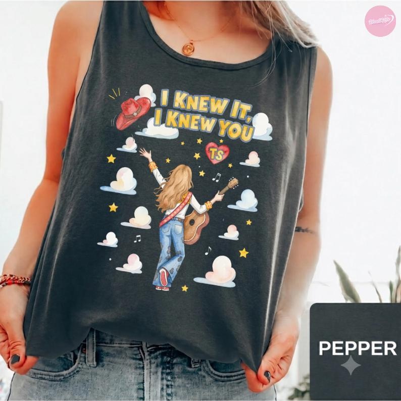 May include: A charcoal gray tank top featuring a graphic of a person with long blonde hair playing an acoustic guitar. The design includes a red cowboy hat, white clouds, yellow stars, musical notes, and the text I KNEW IT, I KNEW YOU with a heart.