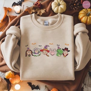 May include: Beige crewneck sweatshirt featuring a Halloween-themed graphic. The graphic includes cartoon characters dressed in costumes, such as a dog in a witch hat, a mummy, a cowgirl, a pig, a dinosaur, and a cowboy. Small bats are also included.