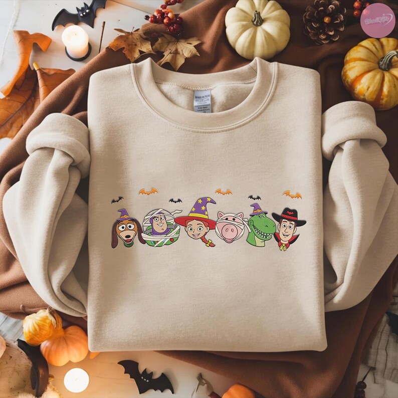 May include: Beige crewneck sweatshirt featuring a Halloween-themed graphic. The graphic includes cartoon characters dressed in costumes, such as a dog in a witch hat, a mummy, a cowgirl, a pig, a dinosaur, and a cowboy. Small bats are also included.