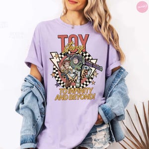 May include: A lilac t-shirt with a "Toy Story" graphic. The design features the words "Toy Story" in red and gold, alongside images of Buzz Lightyear and Woody. The phrase "To Infinity and Beyond!" is also included in the design.