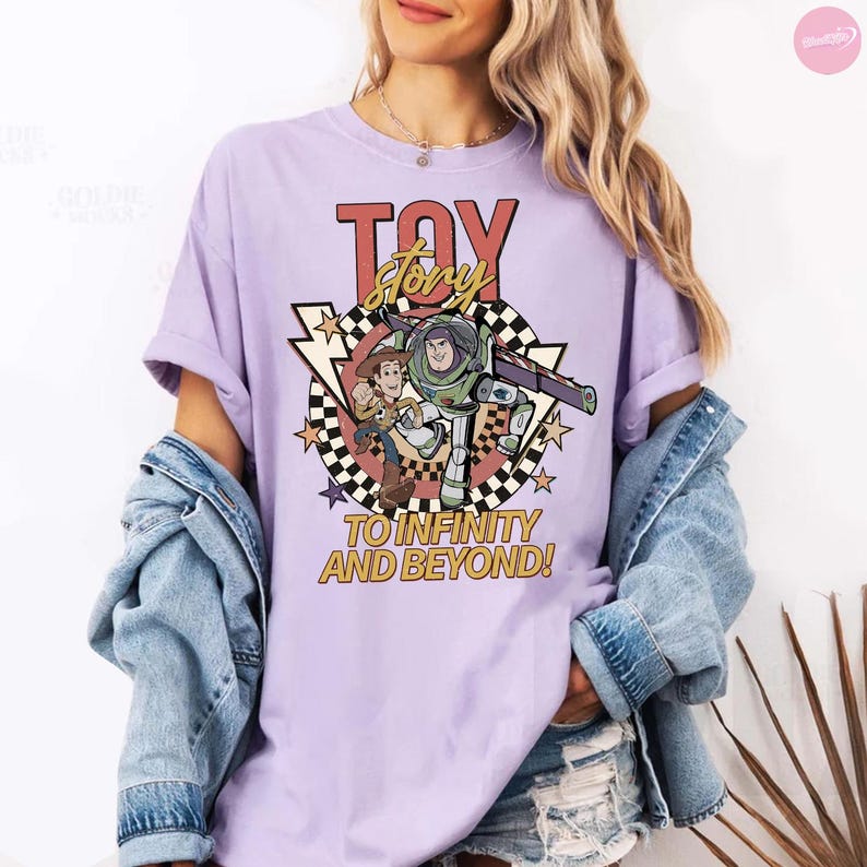 May include: A lilac t-shirt with a "Toy Story" graphic. The design features the words "Toy Story" in red and gold, alongside images of Buzz Lightyear and Woody. The phrase "To Infinity and Beyond!" is also included in the design.