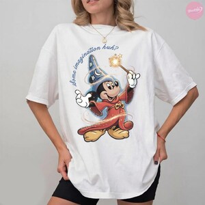 K&ouml;nnte beinhalten: Wei&szlig;es T-Shirt mit einem Cartoon-Mickey-Mouse-Zauberer-Design, der einen Zauberstab mit einem Funkel-Effekt h&auml;lt. Der Text "Some imagination huh?" ist in einer Schreibschrift geschrieben. Das Design verwendet Rot-, Blau- und Gelbt&ouml;ne.