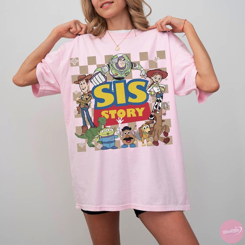 May include: A light pink t-shirt featuring a graphic of characters from the animated film "Toy Story." The design includes the text "SIS STORY" in yellow and red, with images of Woody, Buzz Lightyear, and other characters. The shirt has a checkered background.