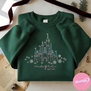 May include: A dark green sweatshirt featuring a castle design with the word "magic" and "EST 1971" in a script font. The castle is outlined in white, pink, and teal. The sweatshirt is folded on a white surface.
