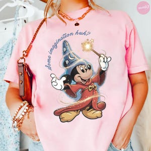 K&ouml;nnte beinhalten: Hellrosa T-Shirt mit einer Grafik von Mickey Mouse als Zauberer, der einen Zauberstab mit einem Stern h&auml;lt. Der Text "Some imagination huh?" ist in Schreibschrift geschrieben. Das T-Shirt hat kurze &Auml;rmel und einen Rundhalsausschnitt.