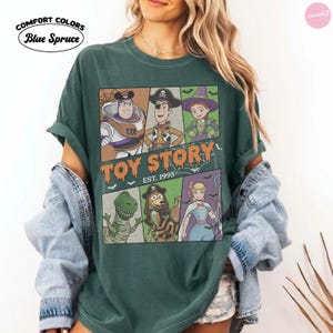 May include: A blue spruce Comfort Colors t-shirt featuring a graphic of Toy Story characters in Halloween costumes. The print includes Buzz Lightyear, Woody, Jessie, Rex, Slinky Dog, and Bo Peep with the text Toy Story Est. 1995 in orange lettering.