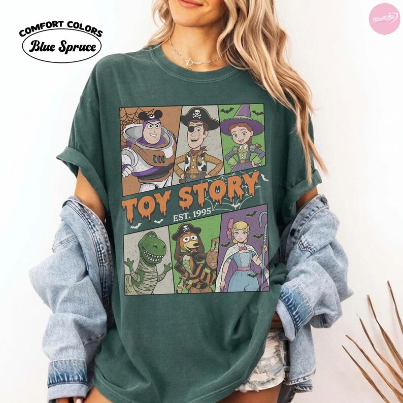 May include: A blue spruce Comfort Colors t-shirt featuring a graphic of Toy Story characters in Halloween costumes. The print includes Buzz Lightyear, Woody, Jessie, Rex, Slinky Dog, and Bo Peep with the text Toy Story Est. 1995 in orange lettering.