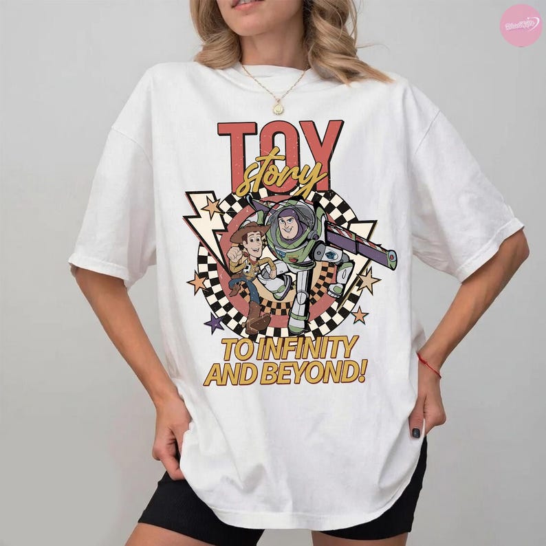 May include: White t-shirt with a graphic from the film "Toy Story." The design features the words "Toy Story" in red and yellow, and images of Woody and Buzz Lightyear. The phrase "To Infinity and Beyond!" is also displayed on the shirt.