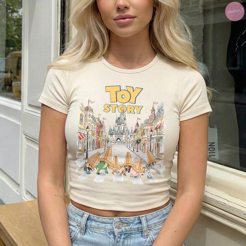 May include: Cream-colored, short-sleeved crop top featuring the words "TOY STORY" in yellow and a colorful graphic of a street scene with characters. The shirt is paired with blue jeans.