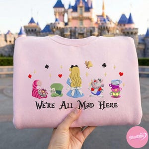 Puede incluir: Sudadera rosa claro con bordados de Alicia en el pa&iacute;s de las maravillas. El dise&ntilde;o incluye el Gato de Cheshire, Alicia, el sombrero del Sombrerero Loco y la frase "We're All Mad Here".