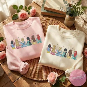 May include: Two crewneck sweatshirts, one light pink and one cream, featuring an embroidered design of cartoon princesses. The sweatshirts are folded on a woven surface, surrounded by roses and books.