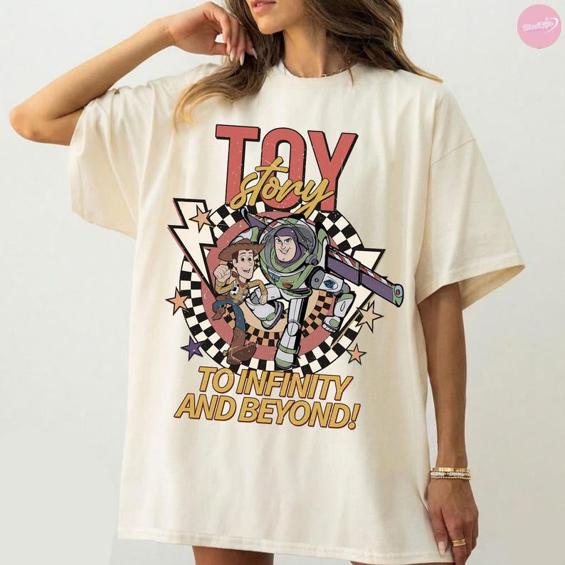 May include: Cream-coloured t-shirt with a graphic from the animated film "Toy Story." The design includes the words "Toy Story" in red and yellow, along with images of Woody and Buzz Lightyear. The phrase "To Infinity and Beyond!" is also displayed.