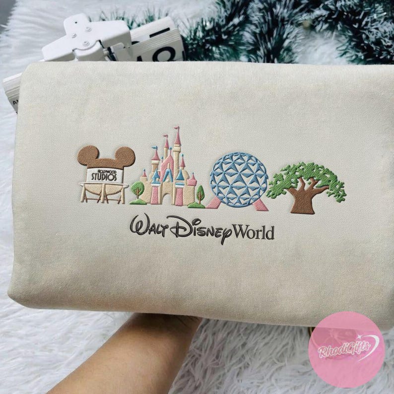 Peut inclure: Pochette beige brod&eacute;e avec le logo Walt Disney World et des images embl&eacute;matiques du parc. Le design comprend le panneau Hollywood Studios, un ch&acirc;teau, Spaceship Earth et un arbre. Le texte "Walt Disney World" est brod&eacute; en dessous.