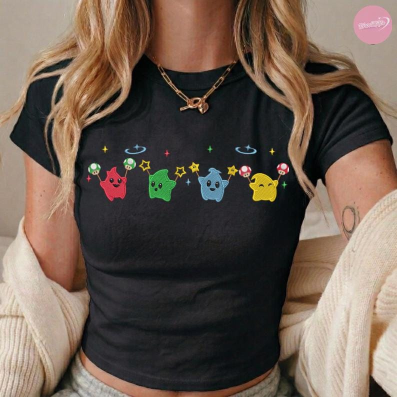 Puede incluir: Top corto negro con un dise&ntilde;o bordado de cuatro personajes de dibujos animados en rojo, verde, azul y amarillo. Cada personaje sostiene una seta y est&aacute; conectado por estrellas. La camiseta tiene mangas cortas y cuello redondo.