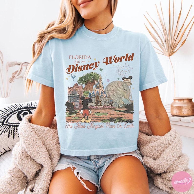 May include: Pale blue t-shirt featuring a retro Disney World design. The graphic includes the text "Florida Disney World" and "The Most Magical Place On Earth" with illustrations of Mickey Mouse and park attractions.
