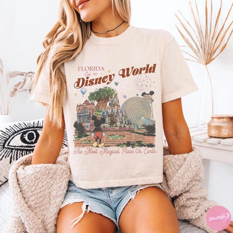 May include: Cream-colored t-shirt featuring a vintage-style graphic of Disney World in Florida. The design includes Mickey Mouse, Cinderella's castle, and the Spaceship Earth, with text that reads "Florida Disney World" and "The Most Magical Place On Earth."