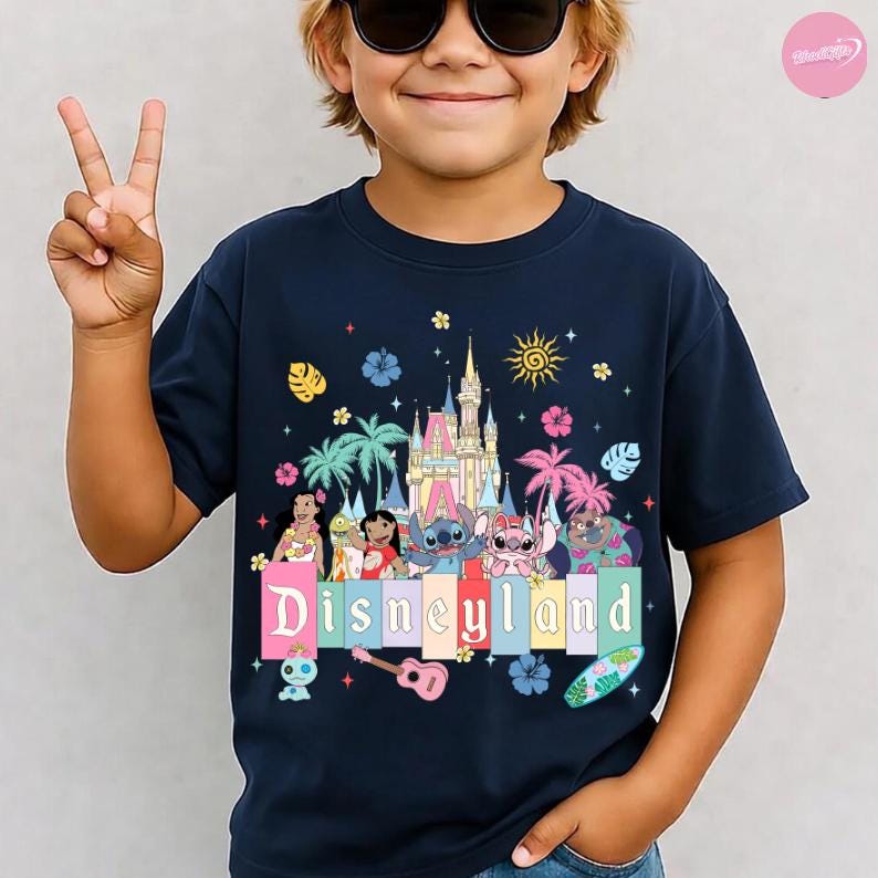 Pu&ograve; includere: T-shirt blu navy con grafica colorata di Disneyland con personaggi dei cartoni animati, un castello e la scritta "Disneyland". Il design include un sole, fiori e palme. La maglietta &egrave; indossata da un bambino.