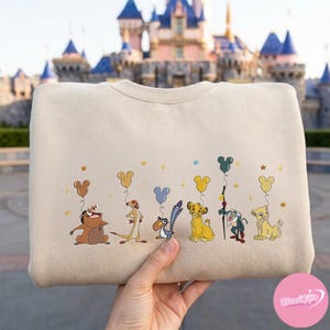 K&ouml;nnte beinhalten: Cremefarbenes Sweatshirt mit einer Illustration von Charakteren aus Der K&ouml;nig der L&ouml;wen, die jeweils einen Mickey Mouse-f&ouml;rmigen Ballon halten. Das Design umfasst Timon, Pumbaa, Zazu, Simba, Rafiki und Nala, mit goldenen Sternen.