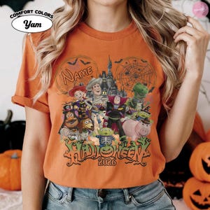 May include: An orange t-shirt featuring a graphic of various animated toy characters dressed in Halloween costumes in front of a castle. The design includes spider webs, bats, the text Name, the word HALLOWEEN, and the year 2026 in black lettering.