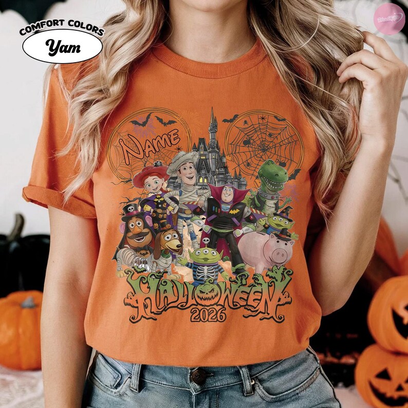 May include: An orange t-shirt featuring a graphic of various animated toy characters dressed in Halloween costumes in front of a castle. The design includes spider webs, bats, the text Name, the word HALLOWEEN, and the year 2026 in black lettering.
