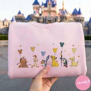 K&ouml;nnte beinhalten: Ein hellrosa Sweatshirt mit Figuren aus Der K&ouml;nig der L&ouml;wen, darunter Timon, Pumbaa und Simba. Das Design umfasst Mickey Mouse-f&ouml;rmige Ballons und Sterne. Das Sweatshirt wird vor einem Schloss pr&auml;sentiert.