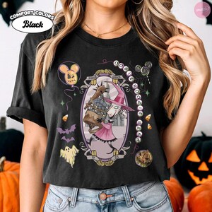 May include: A black t-shirt featuring a graphic print of a character in a pink dress and hat holding a cage. The design includes a balloon, bats, a ghost, candy corn, a lollipop, and text reading OOGIE BOOGIE BASH and 1993 within an oval frame.