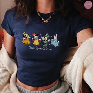 K&ouml;nnte beinhalten: Marineblaues Crop-Top mit gestickten Cartoon-Figuren in bunten Kleidern. Der Satz "Once Upon A Time" steht unter den Figuren. Eine goldene Halskette wird getragen.