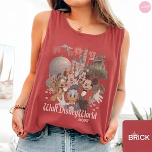 May include: A brick red sleeveless tank top featuring a graphic print of Disney characters including Mickey Mouse, Minnie Mouse, Donald Duck, and Pluto. The design includes the text Walt Disney World Est. 1971 with park landmarks and fireworks.