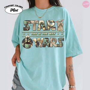 Puede incluir: Camiseta verde menta con el texto "STAR WARS" en un dise&ntilde;o multicolor, con la frase "THIS IS THE WAY" debajo. La camiseta incluye im&aacute;genes de personajes de la franquicia Star Wars, como el Mandaloriano y Grogu.