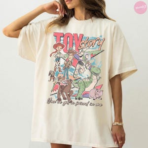 Puede incluir: Camiseta color crema con un gr&aacute;fico colorido de los personajes de Toy Story, incluyendo a Woody, Buzz Lightyear y Rex. El texto "TOY STORY" se muestra encima de los personajes, con la frase "You've got a friend in me" debajo.