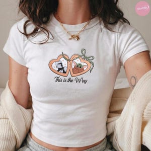 K&ouml;nnte beinhalten: Wei&szlig;es T-Shirt mit einem herzf&ouml;rmigen Design, das eine Figur in R&uuml;stung und einen gr&uuml;nen Au&szlig;erirdischen zeigt. Das Design ist durch eine gr&uuml;ne Schleife verbunden und tr&auml;gt den Text "This is the Way".